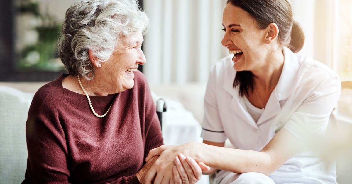 An assisted living nurse and female senior resident holding hands and laughing together